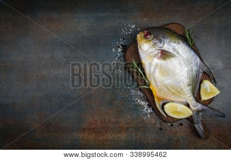 Fresh Pomfret Fish With Herbs Spices Rosemary And Lemon On Wooden Cutting Board And Black Plate Back