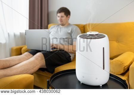 White Air Humidifier Humidifies Dry Air. Man Sitting On Sofa And Working On Laptop. Steam Spraying. 