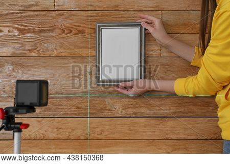 Woman Using Cross Line Laser Level For Hanging Blank Photo Frame On Wooden Wall, Closeup