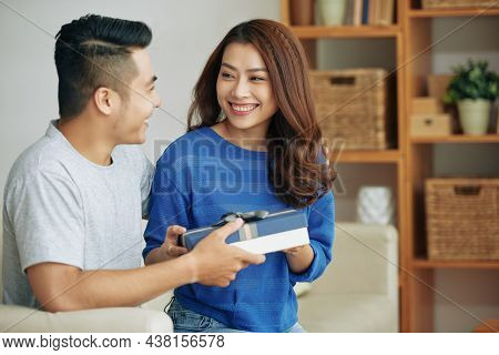 Happy Asian Girl Sitting On Sofa At Home And Receiving Gift From Boyfriend On Anniversery