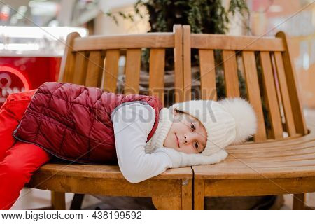 Tired Boy In White Bobble Hat Is Lies With His Mittened Hands Under His Head On A Wooden Bench In Sh