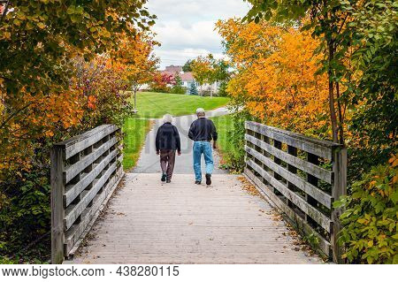 Elderly Couple Walking Over Old Wooden Bridge In Park In Autumn Season. Fall Scenery With Seniors, A