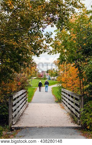 Park In Autumn Season With Elderly Couple Walking Over Old Wooden Bridge. Fall Scenery With Seniors,