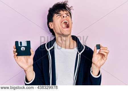 Handsome hipster young man holding floppy disk and sdxc card angry and mad screaming frustrated and furious, shouting with anger looking up. 