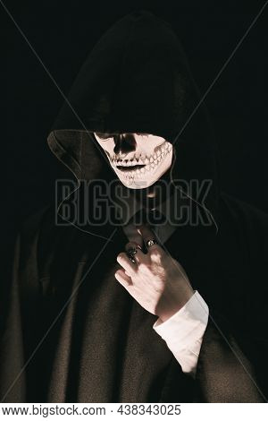 Portrait of a dangerous man hiding his face with skull makeup over the hood of a black robe. Black background. Halloween. Portrait of the devil, death.