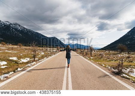 Back View Of Young Woman Walking Along Empty Asphalt Road In The Middle Of Mountainous Area With Rai