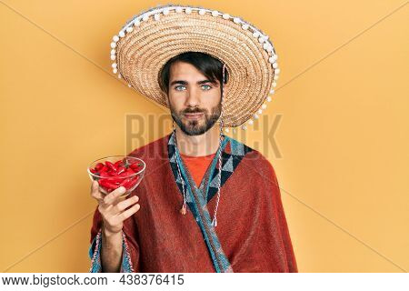 Young hispanic man wearing mexican hat holding chili thinking attitude and sober expression looking self confident 