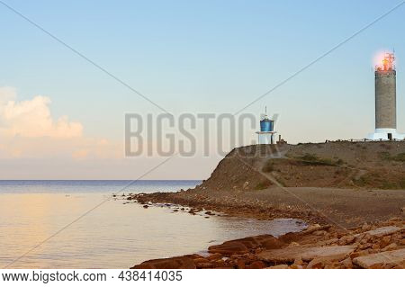 A Tower Lighthouse With A Signal Light On A Steep Seashore In The Rays Of The Rising Sun. There Is A