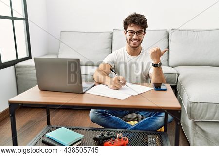 Hispanic man doing papers at home pointing to the back behind with hand and thumbs up, smiling confident 