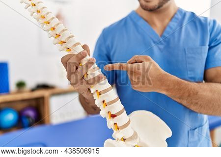 Young hispanic man wearing physio therapist uniform pointing to anatomical model of vertebral column at clinic