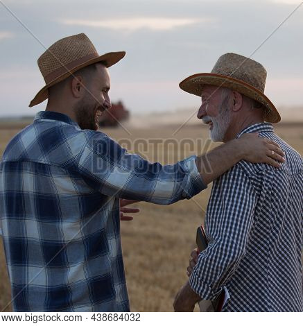 Two Farmers Wearing Plaid Shirts And Straw Hats Talking. Young Agronomist With Hand Of Shoulder Of O