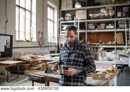 Portrait Of Mature Male Carpenter With Coffee And Smrtphone Indoors In Carpentery Workshop.