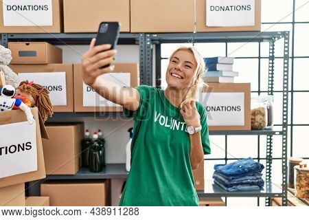 Young blonde volunteer girl smiling happy making selfie by the smartphone at charity center.