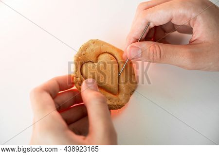 Hands closeup scratch brown sugar caramel candy cookies with a metal needle in the shape of a heart on a white backgound