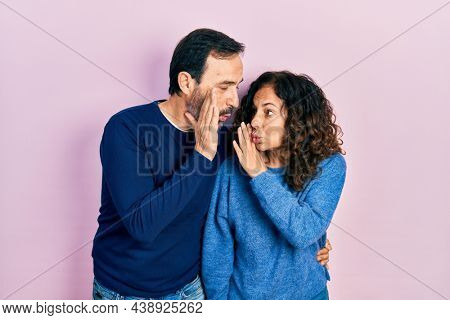 Middle age couple of hispanic woman and man hugging and standing together hand on mouth telling secret rumor, whispering malicious talk conversation 