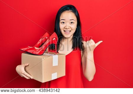 Young chinese girl holding red high heel shoes and package pointing thumb up to the side smiling happy with open mouth 