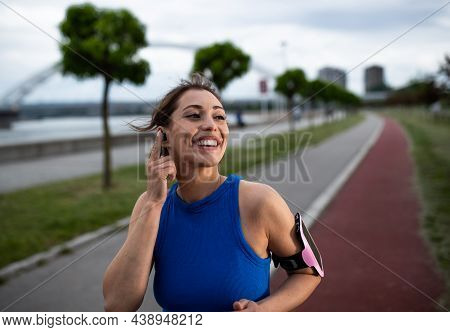 Active Woman Smiling Exerising Outdoors. Young Athlete Using Earbud And Running Jogging Besid Eriver