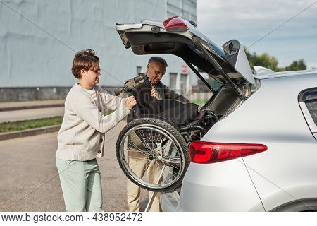Side View Portrait Of Mature Couple Unloading Wheelchair Out Of Car Trunk In Parking Lot Outdoors, C