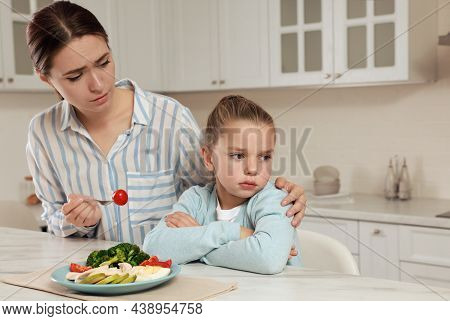 Mother Feeding Her Daughter In Kitchen. Little Girl Refusing To Eat Dinner