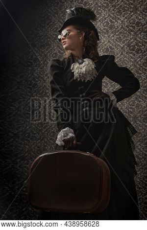 Portrait of an elegant woman in traditional 19th century clothes and round glasses posing on a vintage background with a traveling bag in her hands. Style of the late 19th - early 20th century.