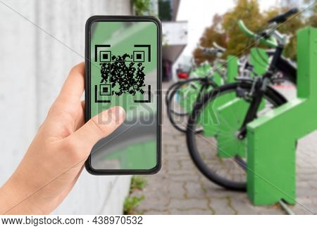 technology, transport and sustainability concept - close up of hand holding smartphone with qr code on screen over bicycles at electric bike charging station on city street on background