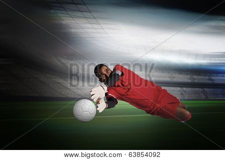 Goalkeeper in red making a save in a large football stadium with lights