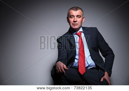Middle aged business man sitting on a stool, resting his hand on his knee while looking at the camera.
