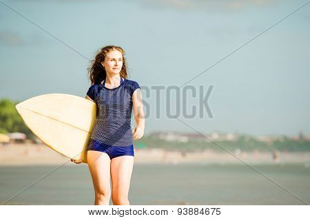 Beautiful sexy surfer girl on the beach at sunset walks along ocean shore, yellow surfboard in her h
