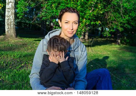 Mother and son sitting on the grass in the park. Family enjoying the outdoors. The kid in the arms of her mother. A boy covers his face with his hands. Shy. Sunny summer evening. A green lawn and trees.