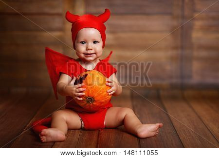 funny baby in devil halloween costume with pumpkin on a dark wooden background