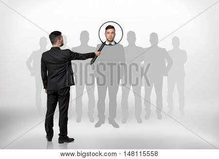 Back view of a businessman looking through magnifying glass on the head of the man that is surrounded by grey silhouettes. Hiring and giving a job. Employment and occupation.