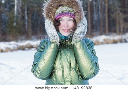 Happy young woman is holding woolly mittens near her head in winter forest outdoors