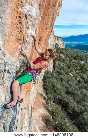 Young Female Climber ascending vertical rocky wall sporty Clothing Red Shirt Green Pants using Rope and other Safety Gear