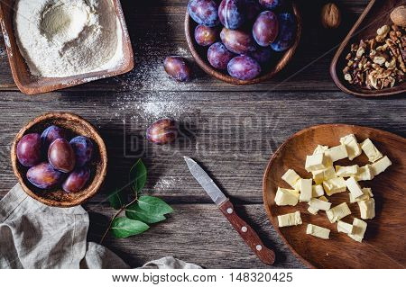 Ingredients for plum pie: fresh plums, walnuts, flour with sugar and chopped butter on rustic wooden table. Overhead view