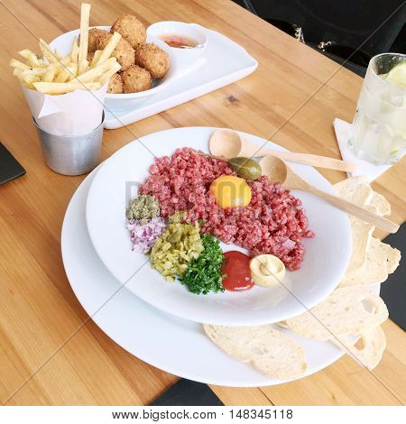 Beautiful beef tartare with egg yolk mustard ketchup scallions red onions cornichons capers and bread on a white plate in a Sao Paulo Brasil restaurant. On table with croquettes fries and caipirinha.