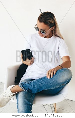 Woman wearing blanc t-shirt, jeans and sneakers sitting on chair and playing with black kitten, toned photo, front tshirt mockup on model, hipster style