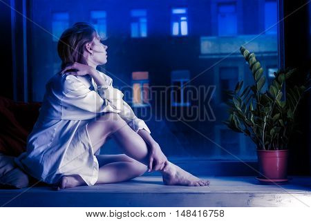 Half-dressed meditative girl in white shirt sits on window sill. Evening urban scene outside the window. Toned in blue colors