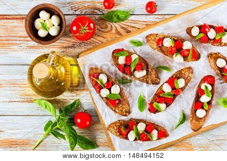 bruschetta with tomatoes mini mozzarella and basil leaves on fried on olive oil baguette with seeds served on chopping board on parchment paper bottle with oil tomato and herbs on wooden background view from above