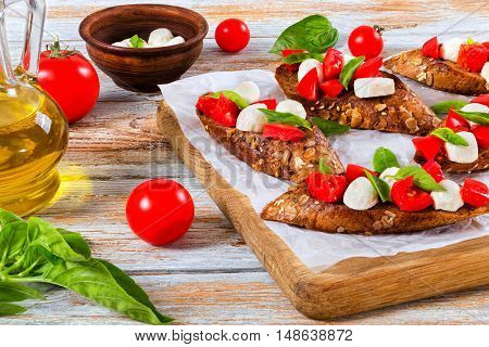 bruschetta with tomatoes mozzarella and basil on fried in olive oil rye baguette with seeds on parchment paper. ingredients on peeling paint white planks italian style close-up