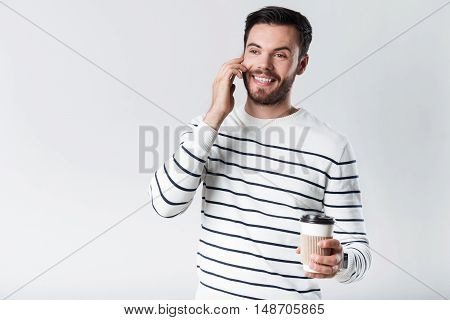 Young handsome smiling man speaking on cellphone and holding coffee while standing against white background.