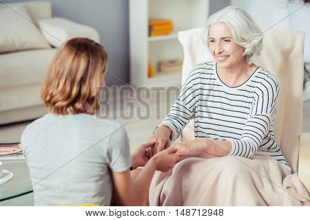 No gap between generations. Loving aged smiling woman sitting in the armchair and expressing joy while holding hands of her adult daughter