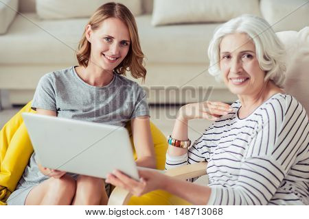 Spend time together. Cheerful smiling aged woman resting with her granddaughter and using laptop while sitting at home