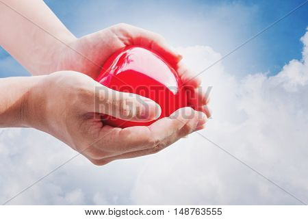 Hand holding red hart on blue sky and white clouds, with bright light
