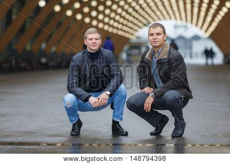 Portrait of two young men sitting on his haunches on background of lights installation main park alley