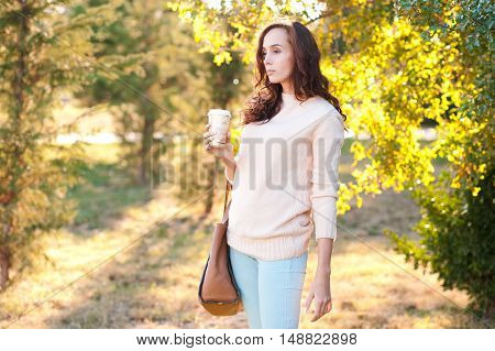 Young girl 20-25 year old wearing casual sweater and denim jeans holding cup of coffee outdoors. Posing over green nature background. .