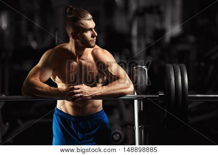 Attractive muscular bodybuilder guy prepare to do exercises with barbell in a gym. Guy bodybuilder tired in gym. Young Man Bodybuilder Resting In Gym After Exercising