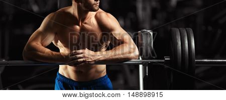 Attractive muscular bodybuilder guy prepare to do exercises with barbell in a gym. Guy bodybuilder tired in gym. Young Man Bodybuilder Resting In Gym After Exercising