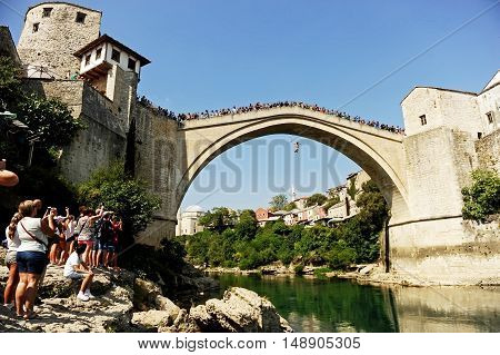 MOSTAR BOSNIA AND HERZEGOVINA - AUGUST 29: Tourists watch a high diver jumping from the old bridge of Mostar into Neretva river on August 29 2015 in Mostar.