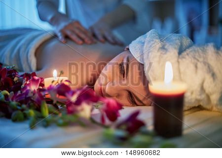 Woman lies on couch with flowers and gets massage lit by two burning candles in beauty salon.