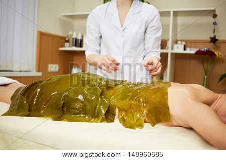 Woman lies on couch with her body algae packed in beauty salon, beautician stands near.